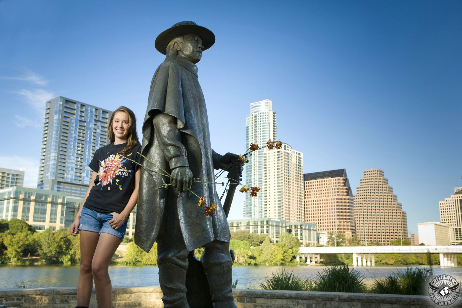 Picture of high school senior girl in Rolling Stones t-shirt leaning against statue of Stevie Ray Vaughn at Auditorium Shores with the Austin skyline behind.